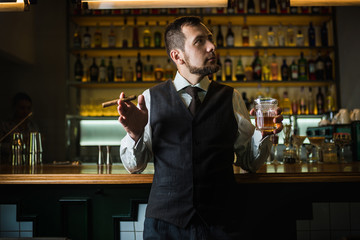 Elegant man in expensive suit near bar counter is smoking cigar and drinking whiskey