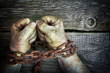 Strong dirty male hands clenched into fists chained with rusty chain on wooden boards background