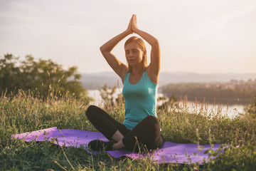 Beautiful sporty woman meditating while sitting on exercise mat with a cityscape and river behinde her.