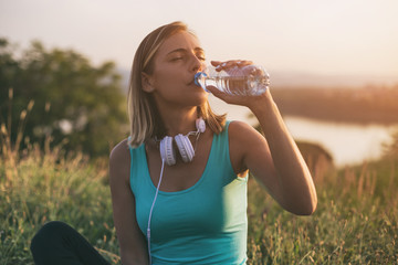 Beautiful sporty woman drinking water while sitting on exercise mat with a cityscape and river...