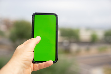 Man holding green screen smart phone with blurred outdoor background.