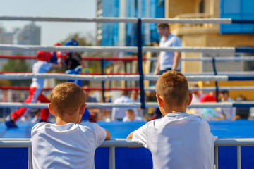 Two boys in white t-shirts watch at the boxing ring with two fight children
