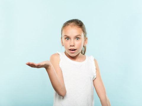 Amazed Cute Girl Holding Virtual Object In Hand. Empty Space For Advertisement. Portrait Of An Astounded Child On Blue Background.