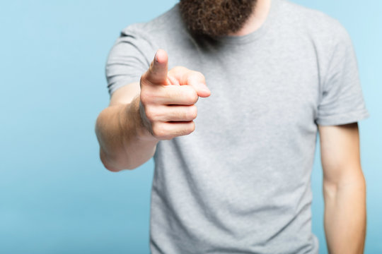 bearded man pointing finger at viewer. indicating gesture. cropped shot of a male torso on blue background. casual hipster in grey t-shirt.