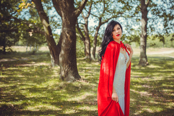 Halloween concept, glamorous costume detail. Young beautiful and mysterious woman in woods, in white Dress and Red Cloak. Little red hood or vampire story

 