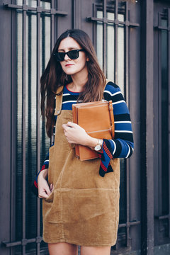 street style portrait of an attractive woman wearing a corduroy short dress, a striped shit, wristwatch and a brown leather bag. fashion outfit perfect for a sunny spring or autumn day