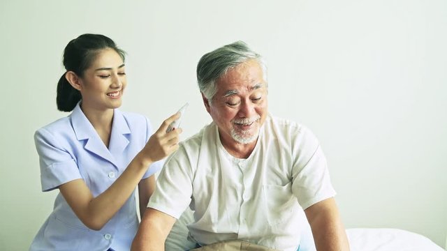 Beautiful nurse using high tech Temperature measurement tool on old man. Senior Asian man with white beard in bed with attractive asian woman nurse. Senior retirement home service, care taker concept.