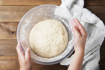 Woman Hands holding a bowl with wheat pizza or pie dough, shaped into ball on floured wooden background. Cooking at home.