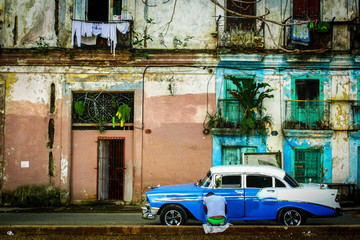 HAVANA, CUBA - DEC 18 2015: A man sits in front of an old classic car near dilapidated buildings next to Parque Cervantes in Havana, Cuba