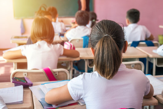 Group Of School Kids Sitting And Listening To Teacher In Classroom From Back