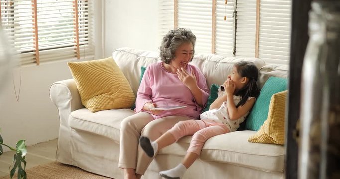 Asian senior woman and young girl are using a digital tablet, talking and smiling while sitting on couch at home. People with technology concept.