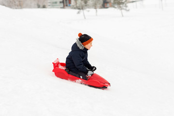 childhood, sledging and season concept - happy little boy sliding on sled down snow hill outdoors in winter