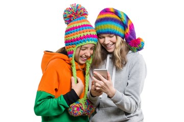 Winter portrait of two happy smiling pretty girls in knitted hats having fun with mobile phone, isolated on white background. People, youth and friendship concept
