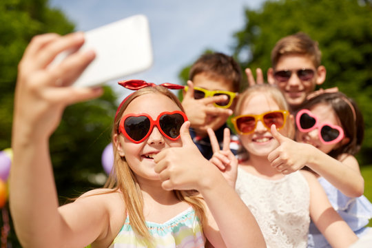 childhood, friendship and technology concept - happy kids in sunglasses taking selfie and showing thumbs up at summer park