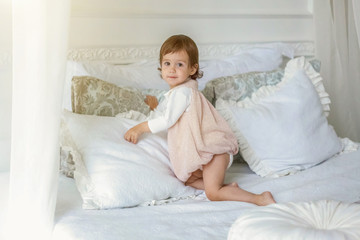 Obraz na płótnie Canvas Funny and cute brunette little smiling girl playing jumping on bed in light bedroom. White interior with big bed. Childhood, preschool, youth, relax concept