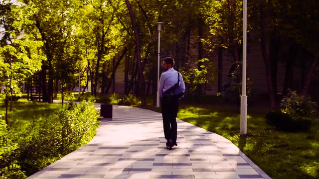 Dolly shot. A man in a business suit riding a kick scooter in a park then he taking scooter and going up stairs. Rear view