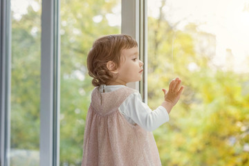 Little cute sweet smiling girl in pastel pink dress standing on the window sill in bright light living room at home and smiling. Childhood, preschool, youth, relax concept