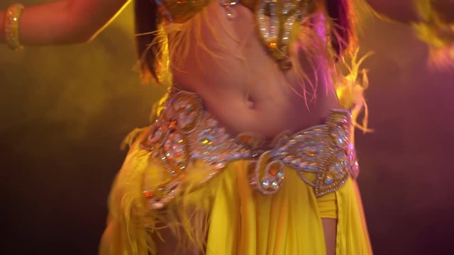 Close up of traditional oriental belly dancer girl dancing on yellow neon smoke background. Unrecognizable woman in exotic costume with feathers sexually moves her hips.