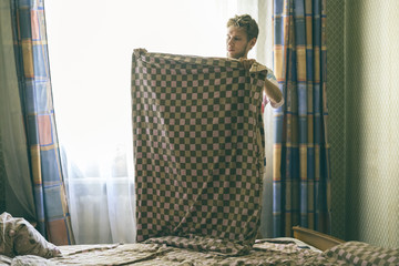 young handsome man in casual makes the bed holding blanket in the morning against the sunlight...