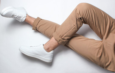Fragment of male legs in beige trousers and white sneakers sits on a white background. Top view. Relaxation. - obrazy, fototapety, plakaty
