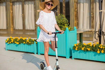 young blonde woman in white dress and hat on blue kick scooter riding on road