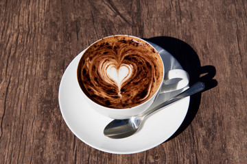 Heart coffee art on a cappuccino in a white cup and silver tespoon on a brown wooden table.