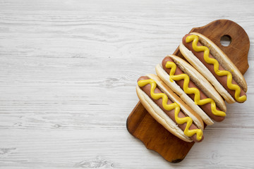 Hot dog with yellow mustard on wooden board on white wooden surface, top view. Space for text.