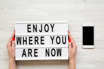 Female hands hold modern board with 'Enjoy where you are now' word, smartphone with blank screen on white wooden background, top view. Business concept. From above, flat-lay, overhead.