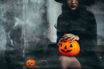Fototapeta premium Young woman in black dress with witch hat and orange pumpkin in hand is on a black background, Halloween concept. Horror theme.