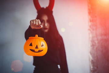 Young woman in black dress with red devil and orange pumpkin in hand is on a black background, Halloween concept. Horror theme.