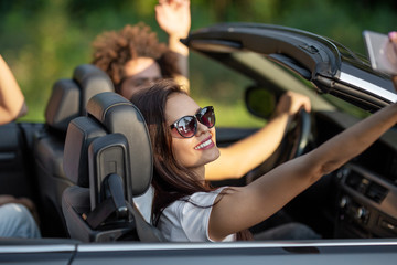 Attractive young brunette woman in sunglasses sits with friends in a black cabriolet smiles and...