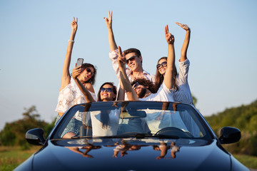 Young lucky girls and boys in sunglasses are sitting in a black cabriolet on the road holding their...