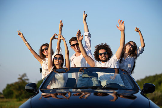 Stylish young girls and guys in sunglasses are sitting in a black cabriolet on the road holding their hands up and smiling on a sunny day.