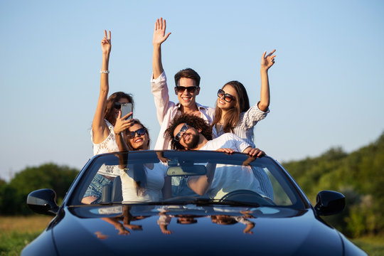 Joyful young girls and guys in sunglasses are sitting in a black cabriolet on the road holding their hands up and smiling on a sunny day.