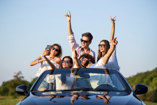 Nice young girls and guys in sunglasses are sitting in a black cabriolet on the road holding their hands up and making selfie on a sunny day.