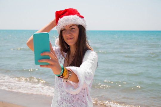 Christmas young smiling woman in red santa hat taking picture self portrait on smartphone at beach over sea background. toned