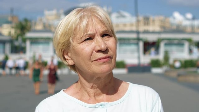 Portrait of blond smiling woman standing outdoors looking at camera. Female pensioner traveling in Moscow, Russia. People walking around. Hand-held camera