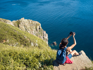 Naklejka premium Young woman photographer sitting on the edge of cliff and taking a photo of landscape. Beautiful rocky shore