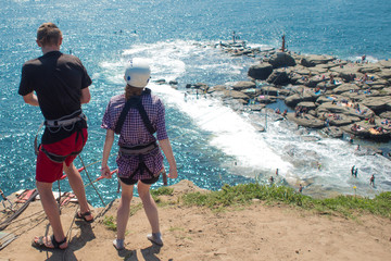 Two are preparing to jump with a bungee from a cliff above the sea, back view,