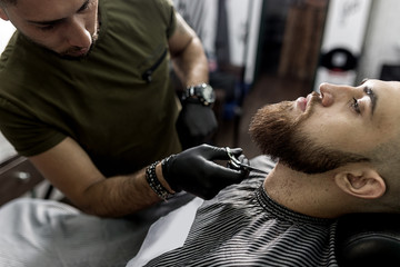 Stylish man with a beard sits at a barbershop. Barber trims mens beard with scissors.