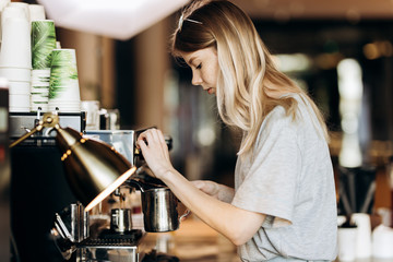 A youthful pretty thin blonde with long hair,dressed in casual outfit,is cooking coffee in a modern coffee shop. Process of making coffee is shown.