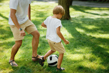 Little blond boy wearing in the white t shirt and shorts plaing footboll with his father on the green lawn in the open air.