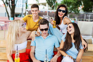 A company of good-looking friends wearing sunglasses laughing and drinking yellow cocktails and socialising at the table in the nice summer cafe. Cheers. Entertainment, having good time. Friendship.