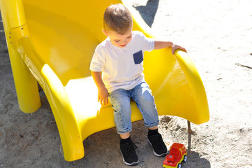 A cute two-year-old boy in a white t shirt sliding down on a yellow slide on a playground