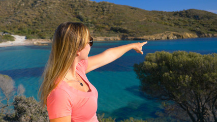 Fototapeta na wymiar CLOSE UP: Unknown young woman points towards the scenic beach from a viewpoint.