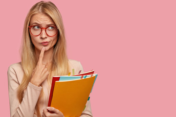 Horizontal view of thoughtful schoolgirl purses lips, keeps index finger on chin, wears round...