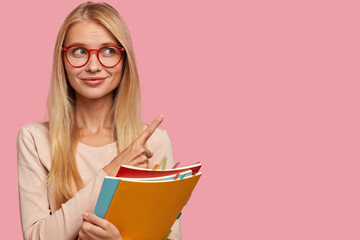 Cheerful pleasant looking European woman with light hair, wears glasses, points with index finger at upper right corner, shows free space for your advertisement, carries textbooks for studying