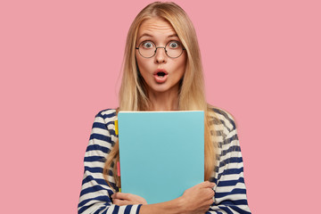 Stupefied light haired Caucasian woman with bugged eyes, bated breath, holds blue folder near...