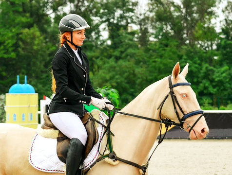 Beautiful girl and horse in jumping show, equestrian sports. Isabelline Akhal-Teke mare with blue eyes and pretty redhead woman in uniform going to jump.