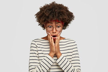 Emotional surprised African American woman with stupefied expression, keeps both hands on cheeks,...
